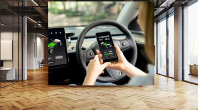 Young woman inside the vehicle, checking EV vehicle's application on battery recharging electricity status display on smartphone screen in modern EV car on her road trip journey. Exalt Wall mural