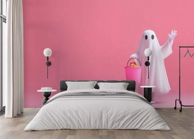 A child dressed as a ghost holds out a pink bucket for candy, waving against a pink background, with ample space for copy space. Wall mural