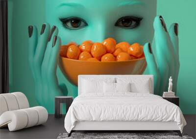 A freaky head made of teal with black eyes and two hands holding a bowl full of orange candy, on a pastel green background.Minimal creative Halloween holiday food concept.Copy space,flat lay  Wall mural