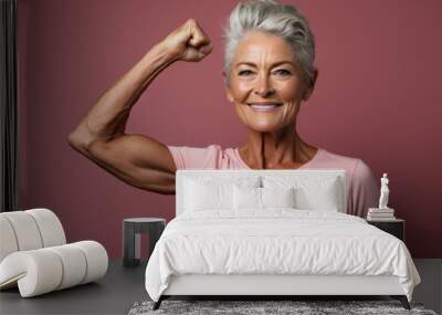 Beautiful cheerful middle aged senior woman with healthy lifestyle, smiling and flexing arm muscles on pink background, health and wellness for aging society concept. Wall mural