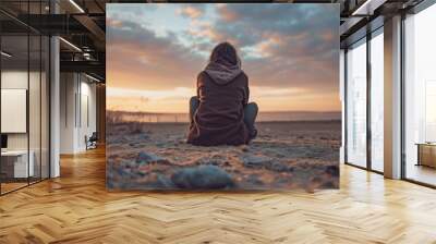 A woman sits on the beach at sunset, looking out at the water Wall mural