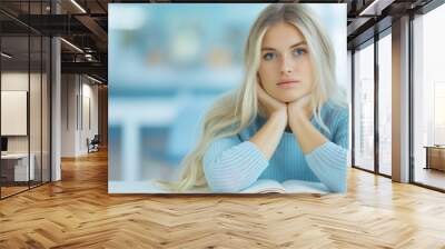 A beautiful blonde woman with blue eyes sitting at a table and thinking about something, an open book on her desk, with a blurry background of a modern university classroom. Wall mural