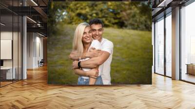 Cute loving couple standing on grass in nature green park with beautiful sundown light. Looking camera hugging. Wall mural