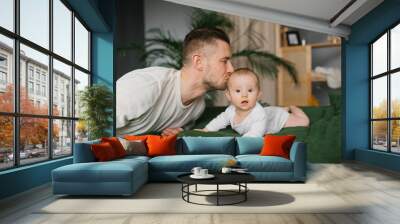 A happy cheerful father sits next to the bed on which his little son is lying and kisses him on the head Wall mural