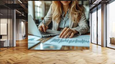 Office worker woman is sitting at a desk with a laptop and a stack of papers and writing on the papers with a pen, working on a project or a report Wall mural