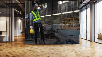 African American male worker wearing vest and helmet safety feeling tired from working hard after work by using shovel to scoop coal on floor for burning at heavy metal industrial factory. Wall mural