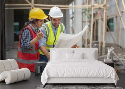 Asian female civil engineer and caucasian male architect wears safety vest with helmet discuss and look at blueprints in housing estate construction site Wall mural