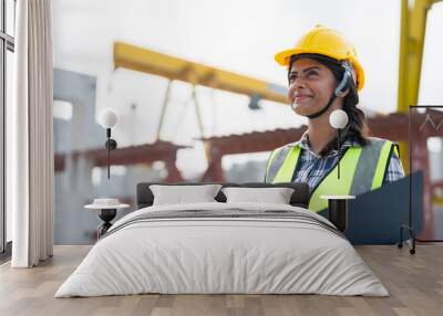 Asian woman engineer holding document smiling at construction site. Confident female Indian wearing protective helmet and vest working in factory making precast concrete wall for real estate housing. Wall mural