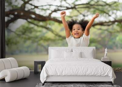 Happy African american little girl smiling and raises her hand while sitting in the park Wall mural