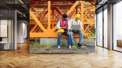 Two construction workers wearing safety vests and hard hats discuss project plans on a laptop at the factory for assembly tower crane. Wall mural