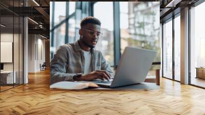 A handsome man in glasses is sitting at the table and working on his laptop, holding an open notebook with notes near him. Wall mural