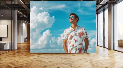 A young man in his thirties, wearing sunglasses and summer attire with floral patterns on the shirt, is smiling while standing at the beach. The background features a blue sky with white clouds, Wall mural