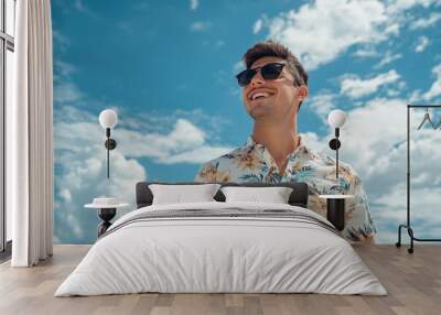 A young man in his thirties, wearing sunglasses and summer attire with floral patterns on the shirt, is smiling while standing at the beach. The background features a blue sky with white clouds Wall mural