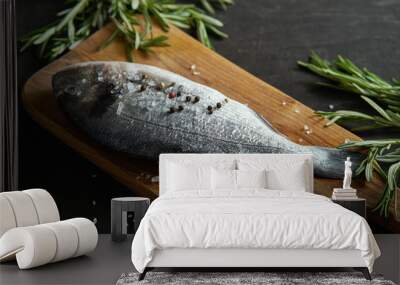 one fresh raw dorada fish with rosemary, pepper and salt on a wooden board and a black table Wall mural