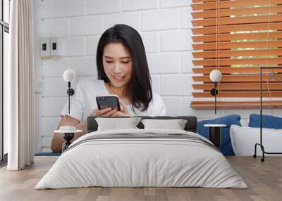 Young asian woman holding a smartphone Play social media, sit on the sofa in the living room at home. Concept of communication technology, doing modern business. Wall mural