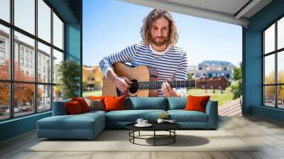 attractive man with long hair with blue eyes and dressed in white and blue striped t-shirt playing acoustic guitar outdoors Wall mural