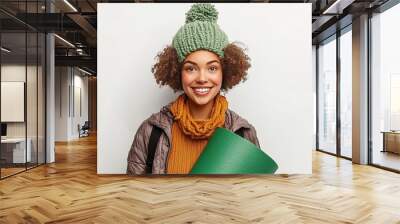 A cheerful young woman wearing a knit hat and scarf, holding a green item, radiating warmth and positivity in a bright setting. Wall mural