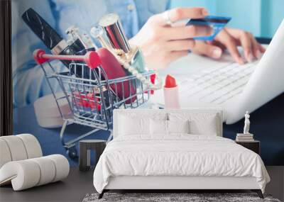 Cosmetics and woman's essentials in shopping cart with woman using laptop and credit card for online shopping Wall mural