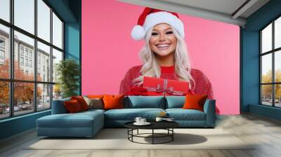 A cheerful person in a Santa hat holds a gift against a pink background, celebrating the holidays. Wall mural