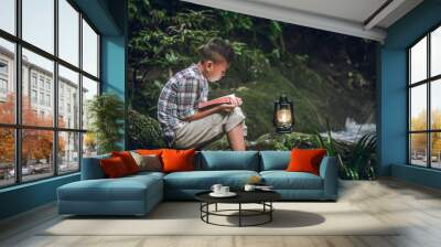 Boy siting on green rock and reading book or bible with oil lamp at waterfall. Children and religion. Wall mural