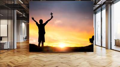 Young man standing holding christian cross for worshipping God at sunset background. christian silhouette concept. Wall mural