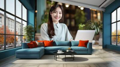 A happy Asian office girl working on her laptop in a cozy cafe, with a blurred background adding to the serene atmosphere. generative Ai. Wall mural