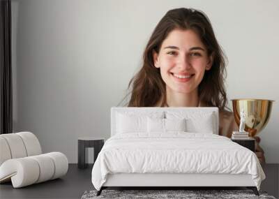 A smiling young caucasian woman in a casual attire holds a gold trophy, her eyes shining with pride. The image captures the essence of victory and the rewards of dedication. Wall mural