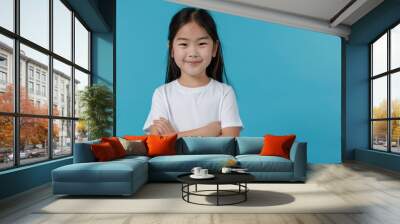 A smiling young girl with dark hair and a white shirt stands against a teal background with her arms crossed. Her bright eyes and happy expression convey a sense of joy and charm. Wall mural