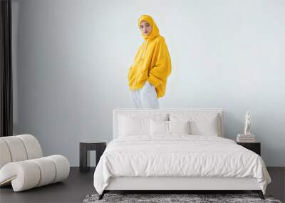 a young Asian woman in a yellow hijab, smiling softly as she poses against a white background, exuding warmth and elegance. Wall mural