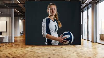 A young female volleyball player stands confidently with a volleyball in her hands. She is dressed in a team uniform and appears focused and ready for the game. Wall mural