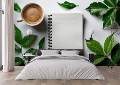 flat lay photograph showcasing blank white spiral notebook resting on table adorned with coffee cups and verdant green leaves, against minimalist white background. Wall mural