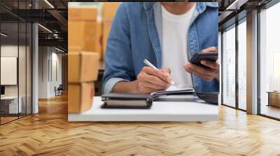 man is busy holding smartphone take order and writing on notebook and parcel box on table and background, online business, small business and e-commerce and delivery service concept Wall mural