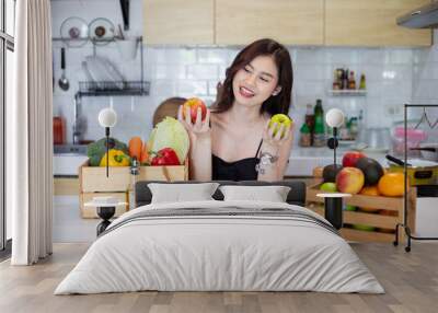 beautiful asian woman smiling preparing, holding a sweet pepper and cut green apple in white kitchen with plenty of fresh vegetables and fruits on the table. At Home Wall mural