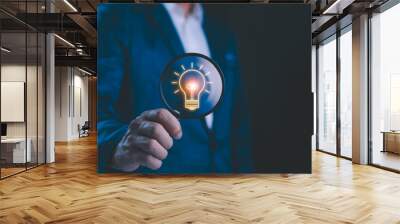 Businessman holding a magnifying glass that illuminates a light bulb icon, representing insight, innovation, and the search for bright ideas in the corporate world. Business solution idea concept. Wall mural