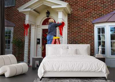 Woman on ladder putting Christmas decorations up on pillars of entrance of two story brick house with bay windows Wall mural