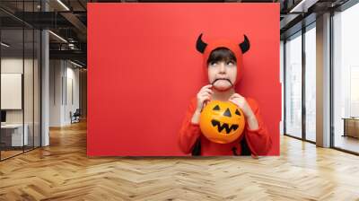 Celebrating Halloween. A boy with a basket of pumpkins in his mouth is dressed in a halloween party devil costume. Studio portrait isolated on red background Wall mural