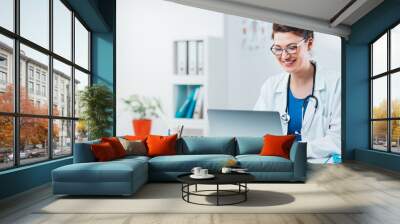 Specialist looking at results from exam on laptop. Friendly woman doctor working on computer in her office Wall mural