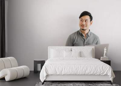 Asian man with beard wear grey shirt typing keyboard working on notebook and holding laptop, happy smiling face looking at empty space and standing isolated over white background wall. Wall mural