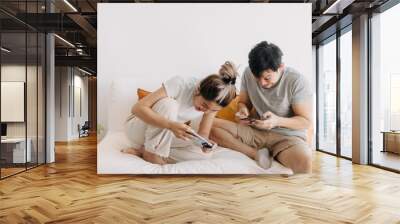 Asian Thai couple using mobile phone, sitting on white sofa in apartment room, both woman and man happy and enjoy playing game online, friend family spending time together. Wall mural