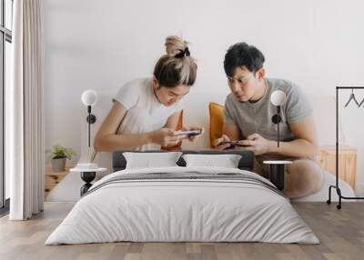 Asian Thai couple using mobile phone together white sitting on white sofa in apartment room, playing game online. Wall mural