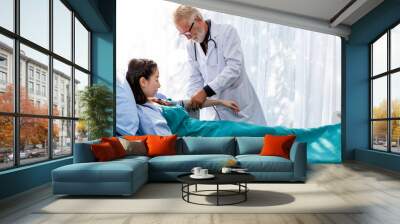 Senior Caucasian professional doctor man check blood pressure with Asian woman patient in the hospital room. Wall mural