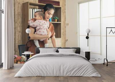 Asia mother stand and working online with a laptop holding her baby daughter at home office. Serious single mom with her child. Asian woman working from home, while in during Covid-19 crisis. Wall mural