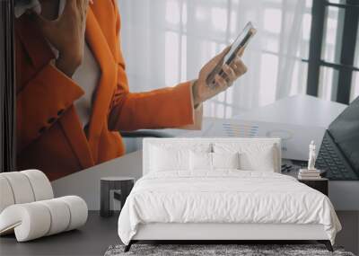 Smiling young Asian businesswoman working on a laptop computer at her desk in a bright modern home office, doing calculating expense financial report finance making notes on paper graph data document. Wall mural