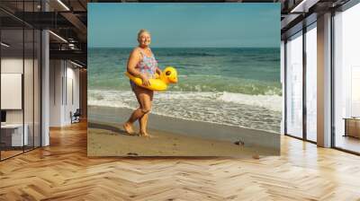 beautiful grandmother 60 plus blonde with a smile on the beach with a lifebuoy yellow duck walks on the sand Wall mural