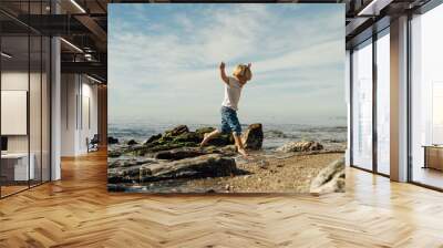 little blond boy playing on the seashore. child playing on the seashore. boy stands on stones in the water. child playing on the seashore Wall mural