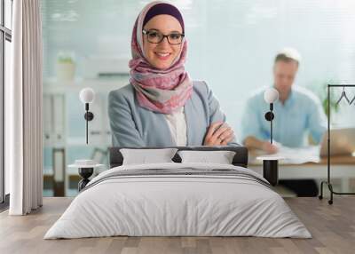 Beautiful young working woman in hijab, suit and eyeglasses standing in office, smiling. Portrait of confident muslim businesswoman. Modern office with big window, man working at desk on background.  Wall mural