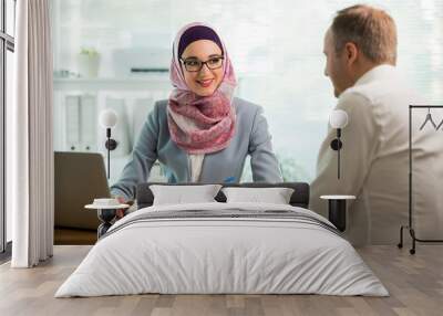 Coworkers meeting in office. Stylish woman in hijab making conversation at desk with man in white modern office. Muslim businesswoman in eyeglasses interviewing man. Wall mural