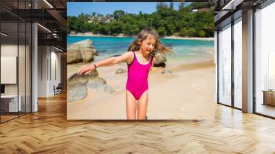 Cute happy little girl running, dancing on the beach in pink swimming suit. Beautiful summer sunny day, turquoise sea, rocks, white sand, picturesque tropic landscape. Phuket, Thailand Wall mural