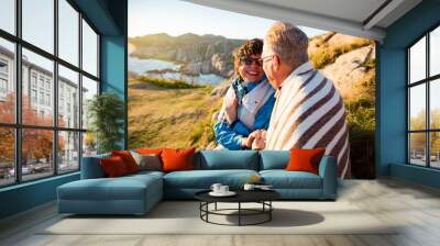 Loving mature couple hiking, sitting on windy top of rock, exploring. Active Mature man and woman wrapped in blanket, hugging and Happily smiling. Scenic view of sea, mountains. Norway, Lindesnes. Wall mural