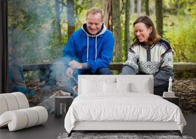 Man and woman roasting on campfire in forest on shore of lake, making a fire, grilling. Happy couple exploring Finland. Scandinavian landscape.  Wall mural